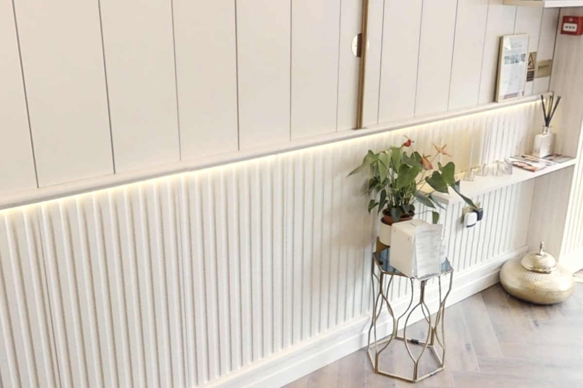 Fluted panelling lit by a warm strip light, with a plant on a brass stand and a herringbone floor, inside Shirland Dental Practice
